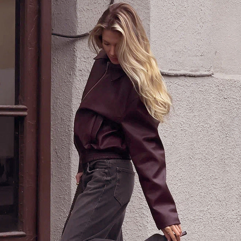 Donna con giacca di pelle marrone scuro e jeans neri, capelli biondi ondulati, cammina in città. Moda autunnale, stile urbano elegante.