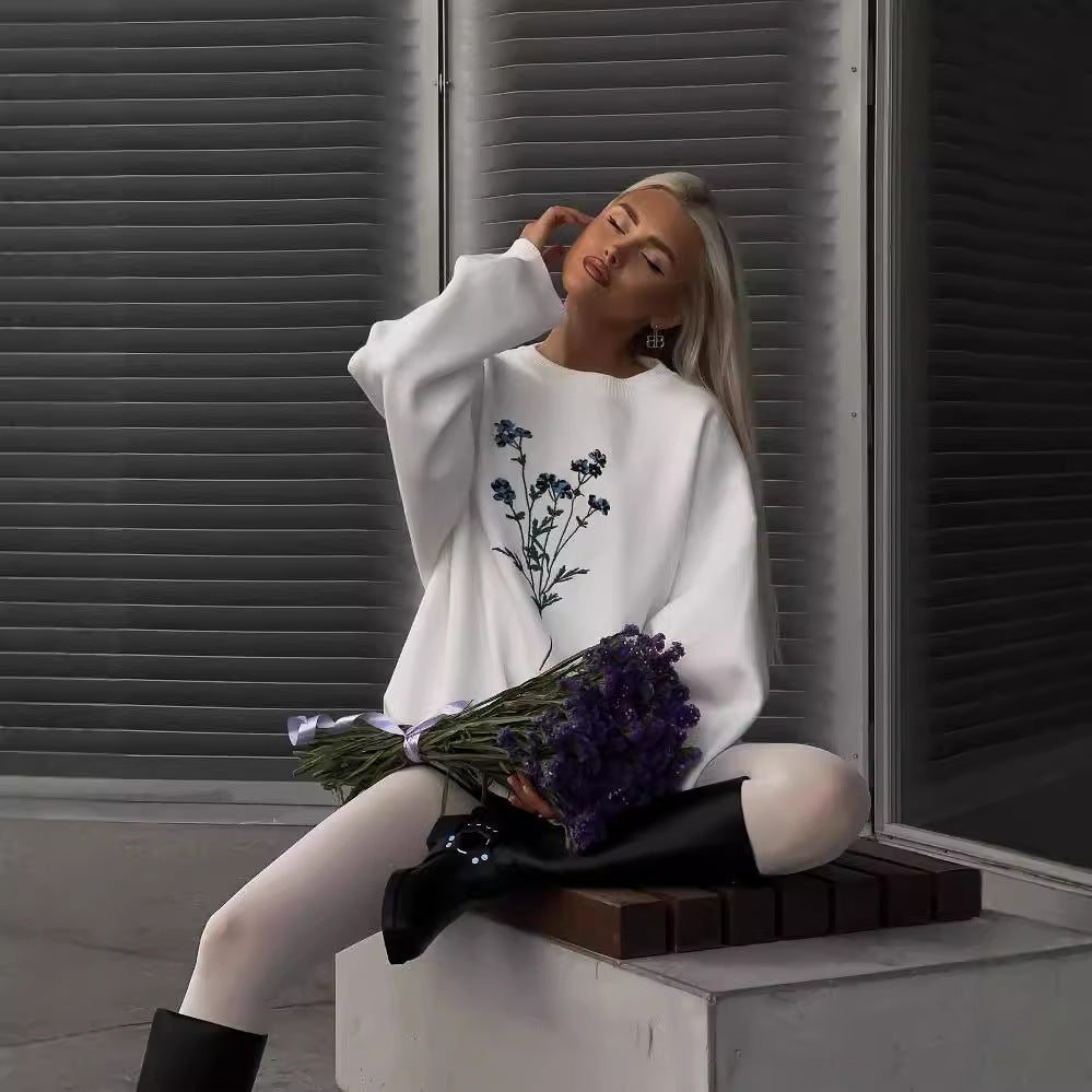 Donna seduta con maglione bianco decorato con fiori blu, tiene un mazzo di fiori viola. Moda autunnale, abbigliamento casual elegante.