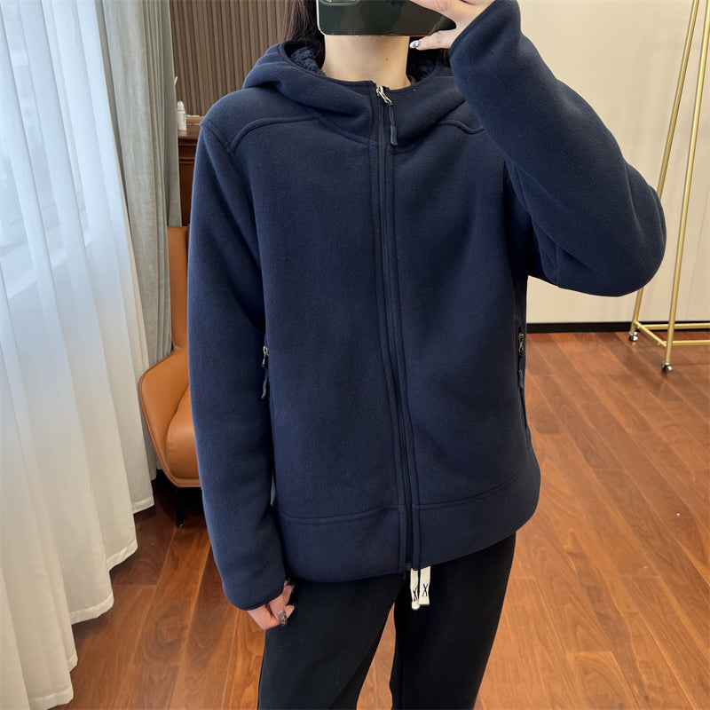 Giacca blu scuro con cappuccio, chiusura a zip, indossata da una persona in piedi su pavimento in legno, vicino a finestra con tende bianche.
