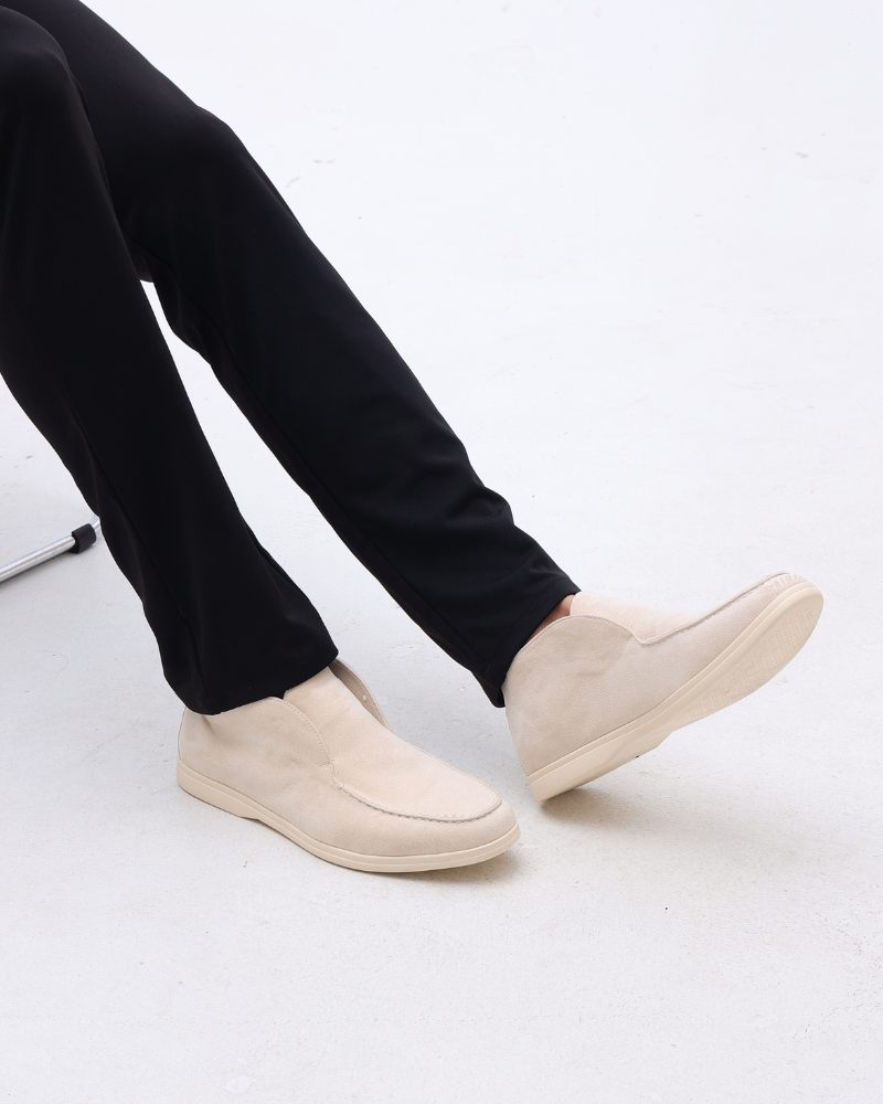 Scarpe mocassino beige su pavimento bianco, indossate con pantaloni neri. Calzature eleganti e comode, ideali per look casual e formali.