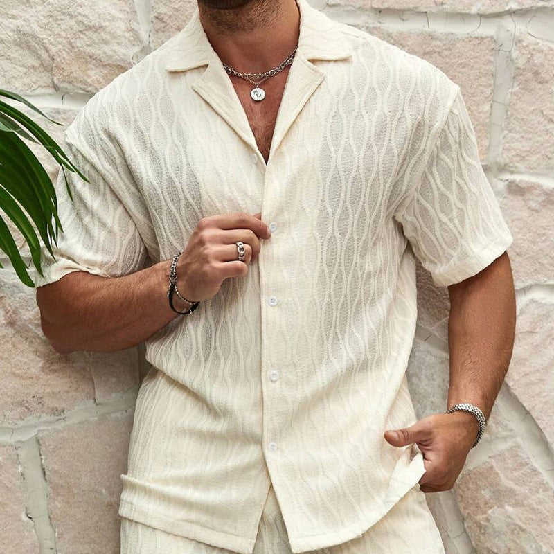 Uomo in camicia estiva bianca a maniche corte con motivo a onde, collana e bracciali, sfondo di muro in pietra chiara. Moda maschile estiva elegante.