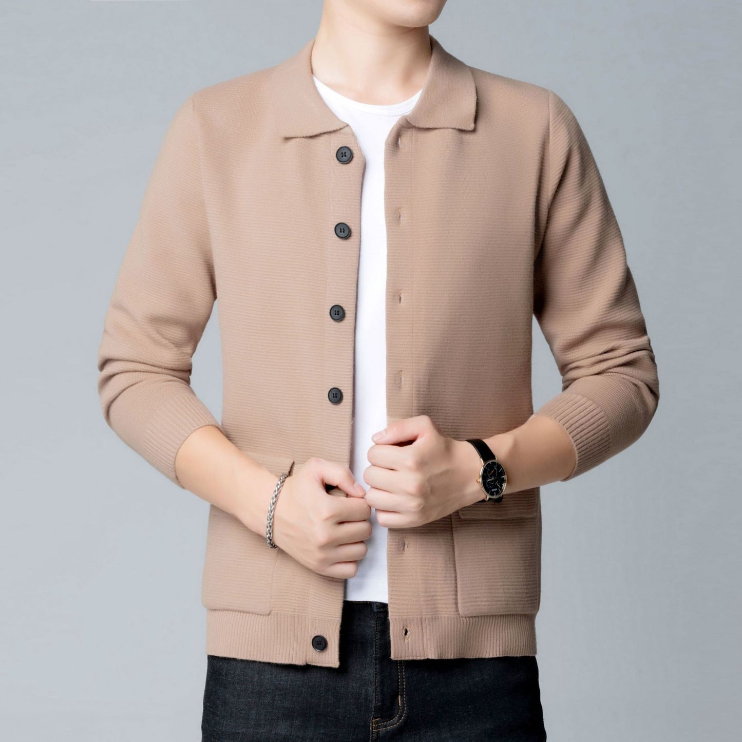 Uomo in cardigan beige con bottoni neri, indossa t-shirt bianca e jeans scuri, orologio nero al polso, sfondo grigio chiaro.