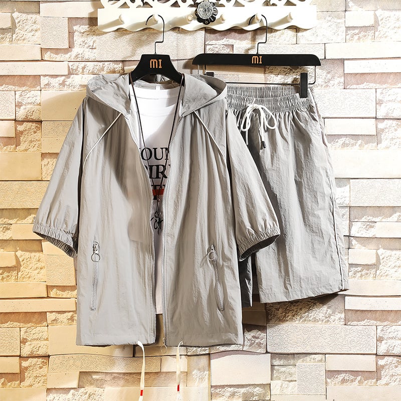 Completo sportivo estivo grigio, giacca con cappuccio e pantaloncini, appeso su grucce. Design casual, ideale per attività all'aperto.