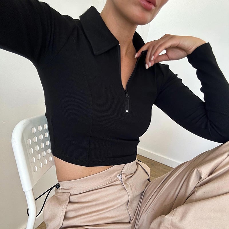 Donna seduta su sedia bianca, indossa maglia nera a maniche lunghe con zip e pantaloni beige. Moda casual, abbigliamento autunnale elegante.