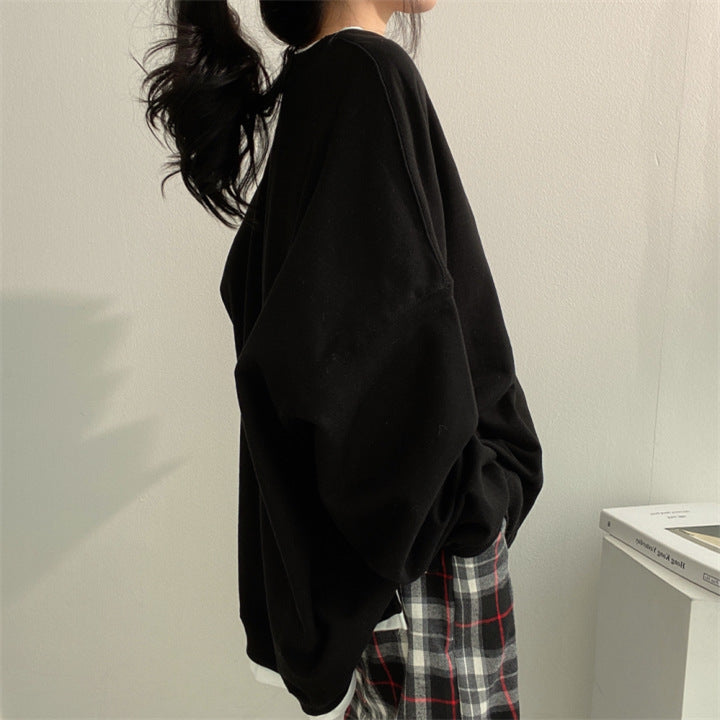 Donna con maglione nero oversize e pantaloni a quadri, capelli raccolti, posa di profilo in interni minimalisti. Moda casual autunnale.