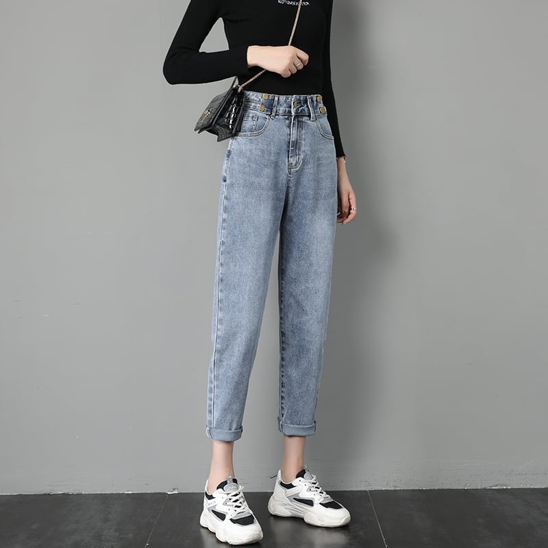 Donna in jeans a vita alta e maglia nera, con borsa a tracolla e sneakers bianche, posa contro un muro grigio. Moda casual femminile.