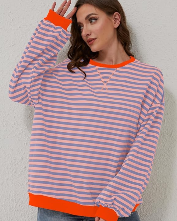 Donna con maglione a righe rosa e blu, bordi arancioni, posa contro parete bianca. Moda casual, abbigliamento autunnale, stile moderno.