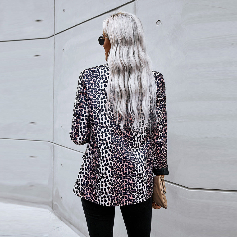 Donna con capelli biondi lunghi indossa giacca leopardata, pantaloni neri e occhiali da sole, cammina su sfondo urbano grigio. Moda autunnale elegante.