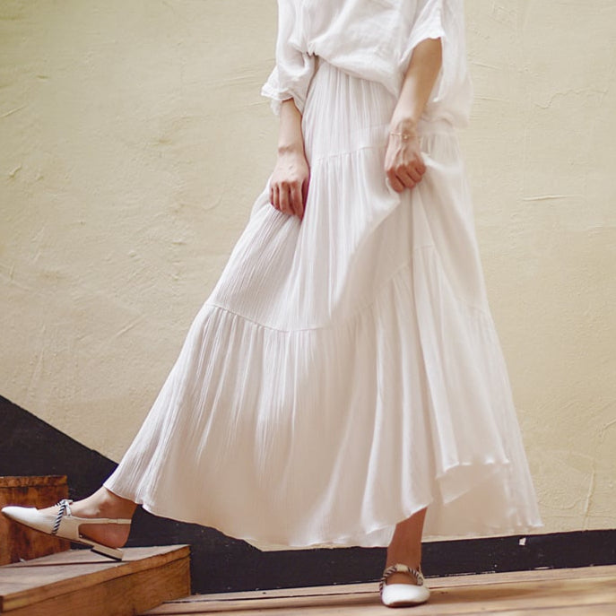 Donna in abito bianco lungo e fluente, scarpe eleganti, posa su scala di legno. Moda estiva, stile boho-chic, abbigliamento femminile elegante.