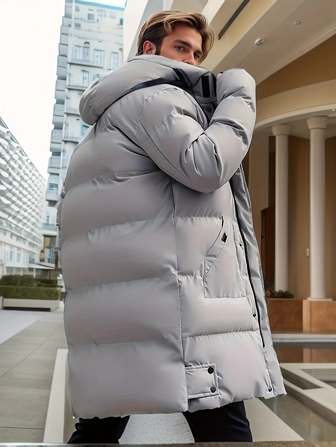 Uomo con cappotto invernale imbottito grigio chiaro, in piedi all'aperto vicino a edifici moderni. Moda maschile, abbigliamento invernale elegante.