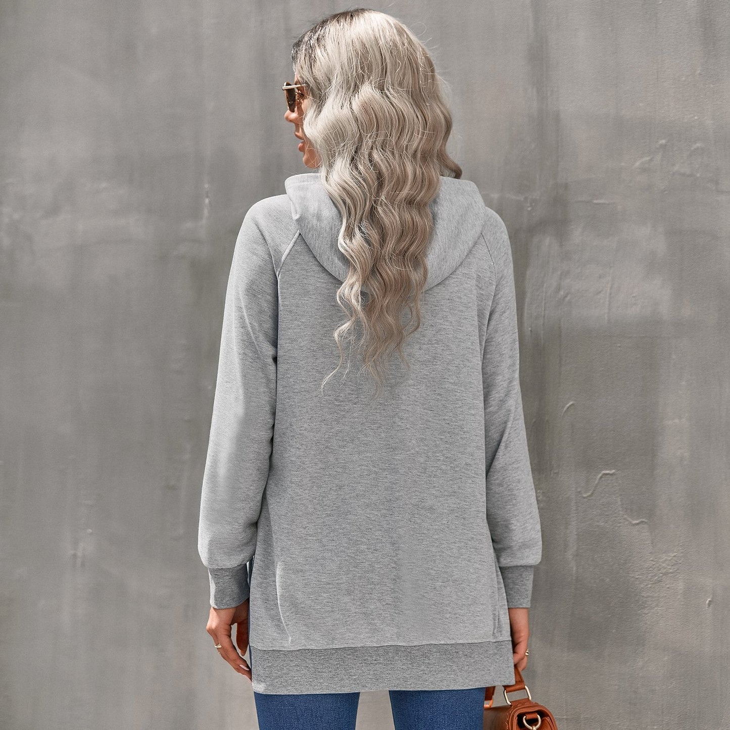 Donna con capelli ondulati indossa felpa grigia con cappuccio, jeans blu, occhiali da sole, sfondo grigio. Moda casual autunnale.