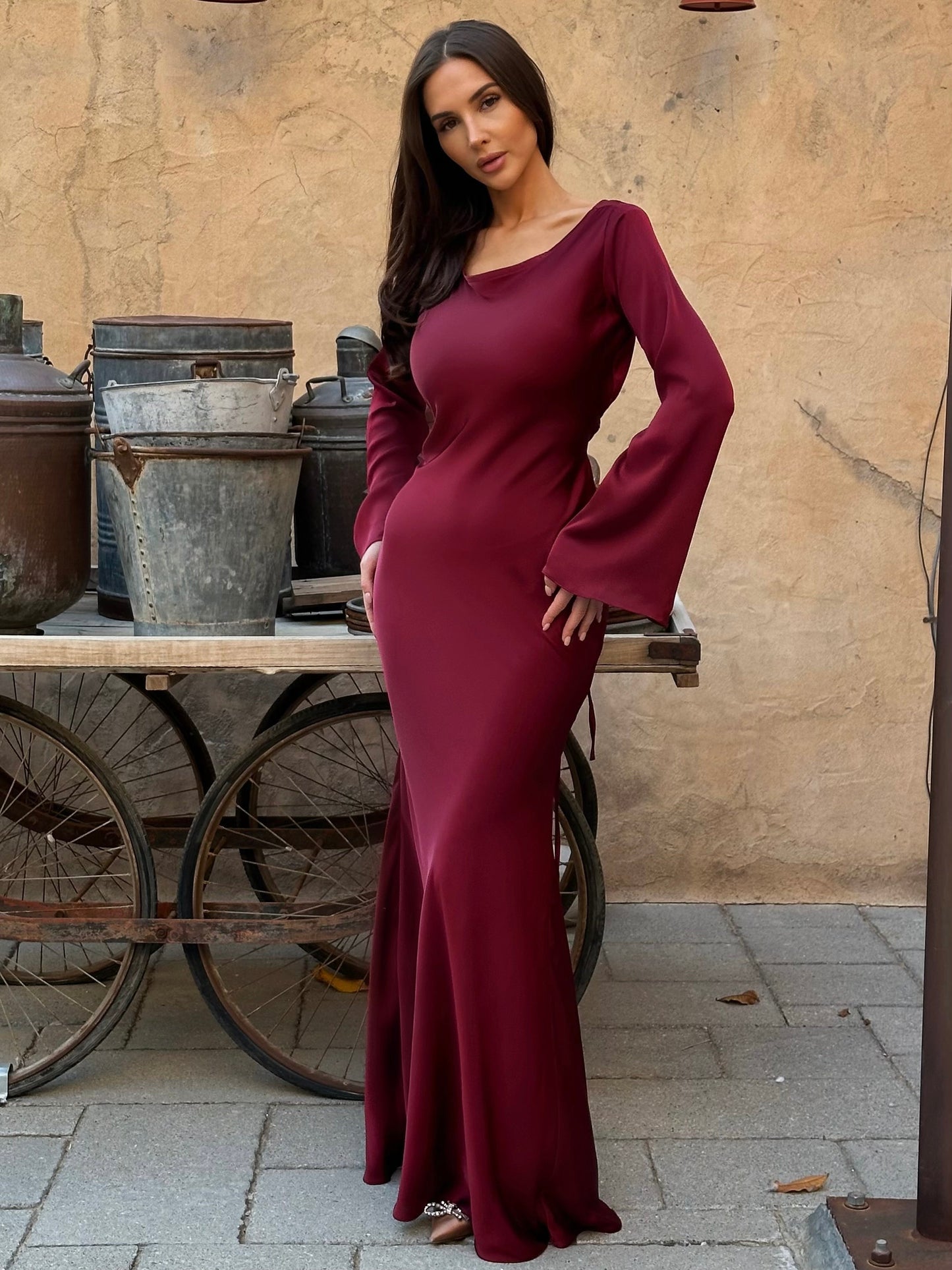 Donna in abito lungo bordeaux elegante, posa davanti a carrelli vintage su sfondo rustico. Moda femminile, abbigliamento elegante, stile sofisticato.