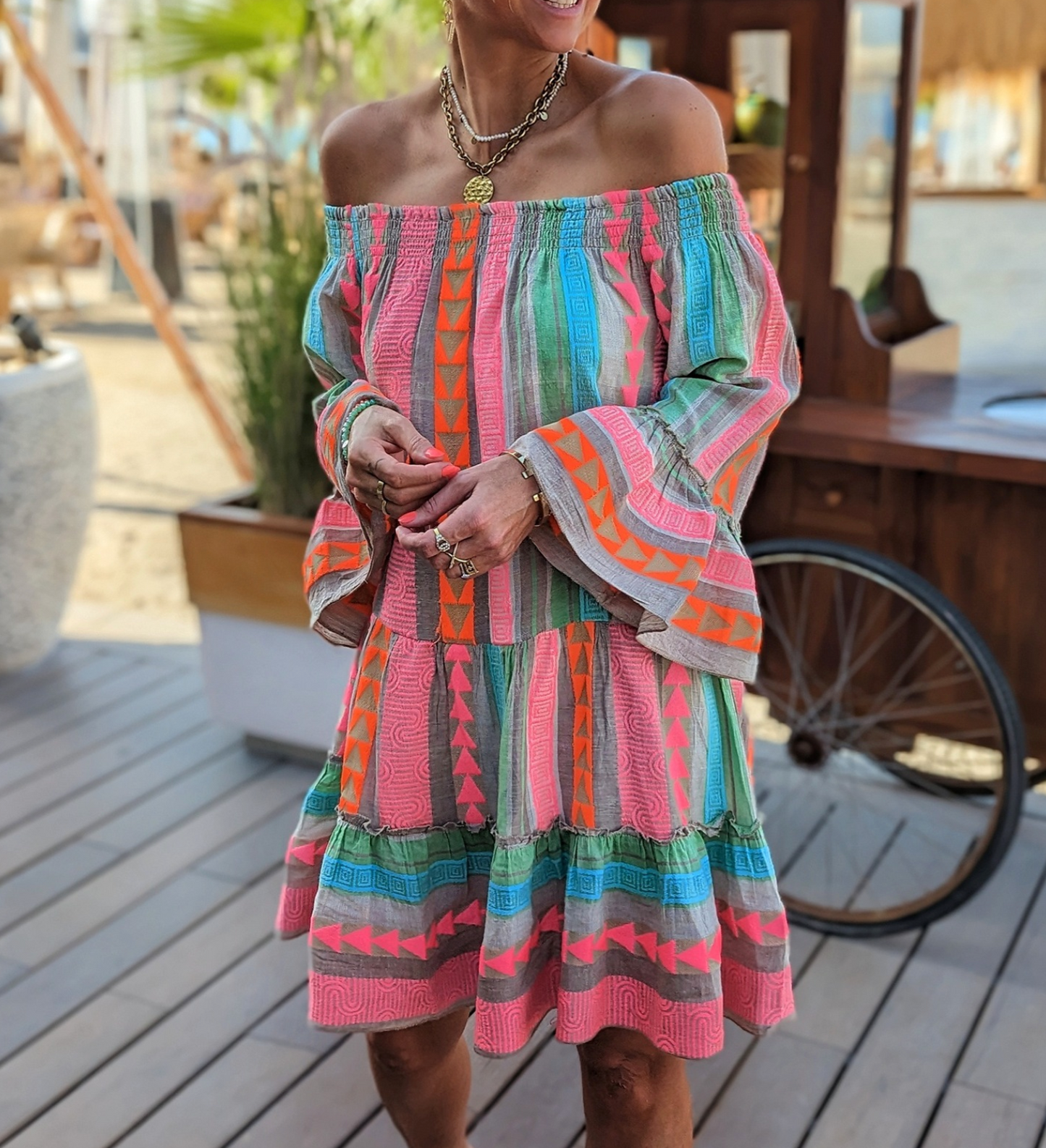 Donna con abito estivo colorato a spalle scoperte, motivi geometrici arancioni e verdi, in un ambiente all'aperto. Moda estiva, abbigliamento casual.