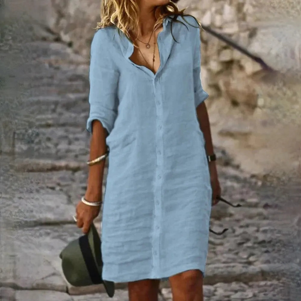 Donna con abito camicia azzurro su strada di pietra, accessori in argento, cappello verde in mano. Moda estiva casual, look elegante e rilassato.