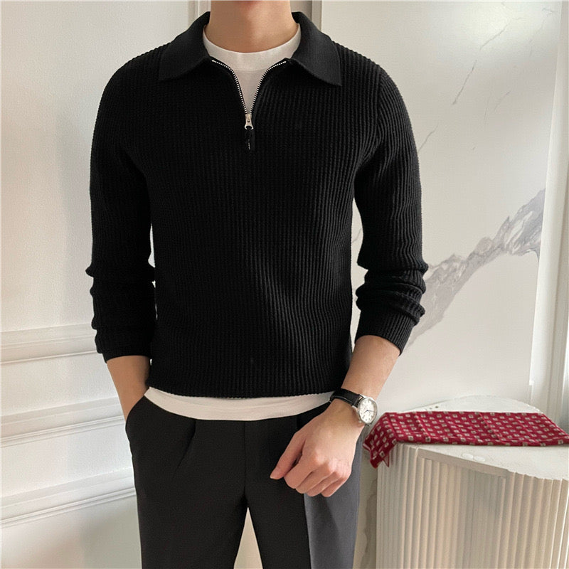 Uomo in maglione nero a coste con zip, camicia bianca sotto, pantaloni grigi. Indossa un orologio elegante. Sfondo interno minimalista.