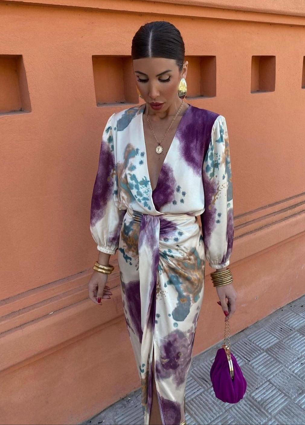 Donna con abito lungo a fantasia astratta viola e beige, borsa viola, orecchini dorati, sfondo arancione. Moda elegante, street style.