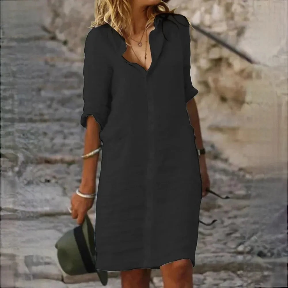 Donna con abito nero casual, maniche lunghe, cammina su strada di pietra. Moda estiva, stile urbano, accessori minimalisti, cappello verde in mano.