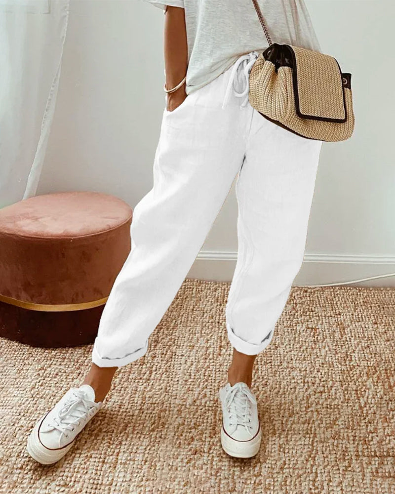Donna con pantaloni bianchi larghi, maglietta grigia, borsa a tracolla beige e sneakers bianche. Moda casual estiva, stile rilassato.