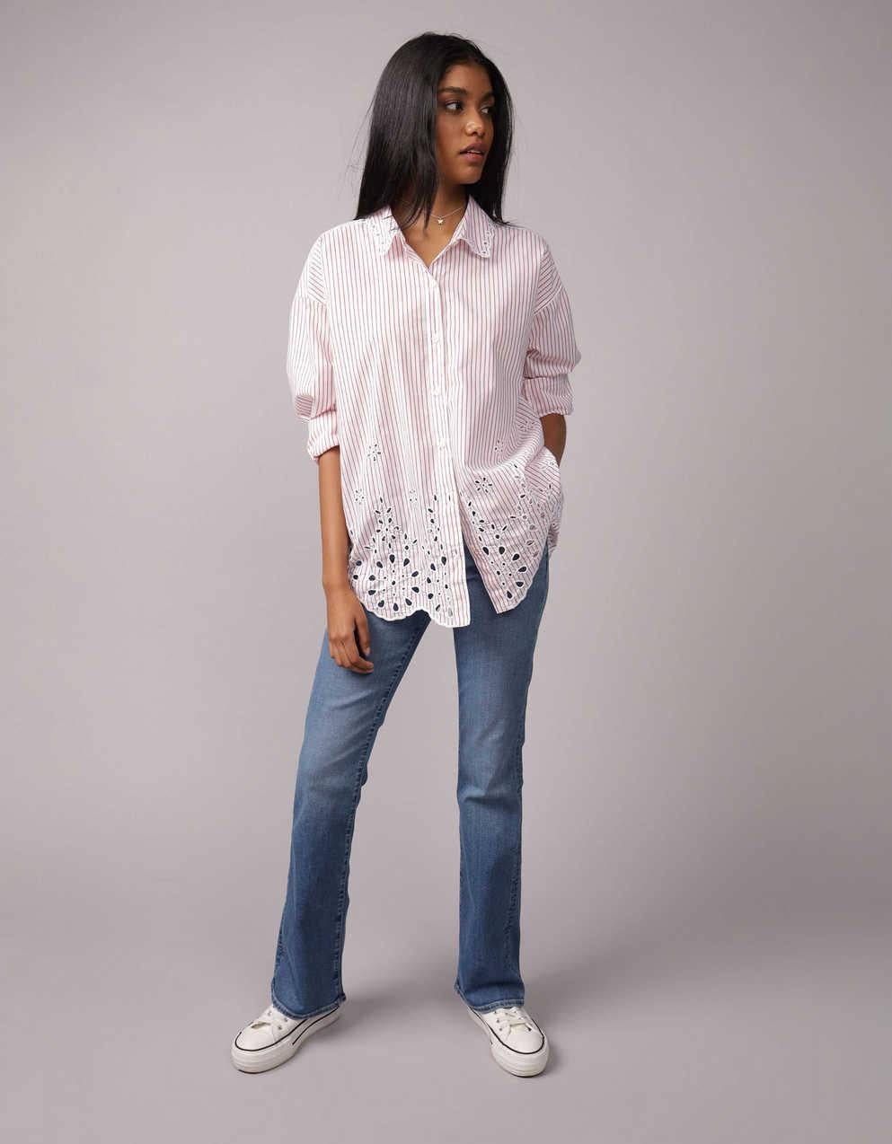 Donna con camicia a righe rosa e jeans a zampa, mani in tasca, posa su sfondo grigio. Moda casual, abbigliamento femminile, stile contemporaneo.