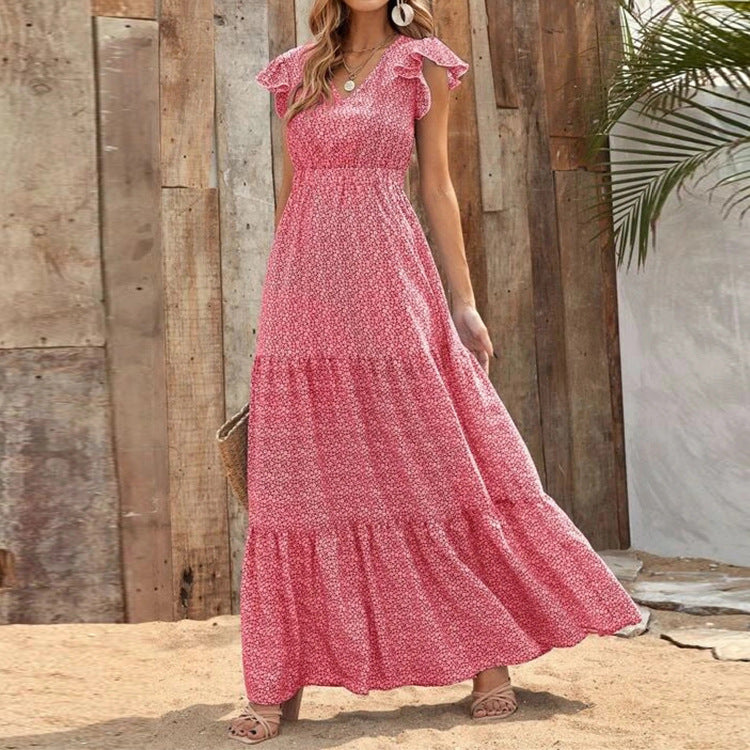 Donna con abito lungo estivo rosa a fiori, in piedi su sabbia, sfondo di legno e foglie di palma. Moda estiva, abbigliamento femminile elegante.
