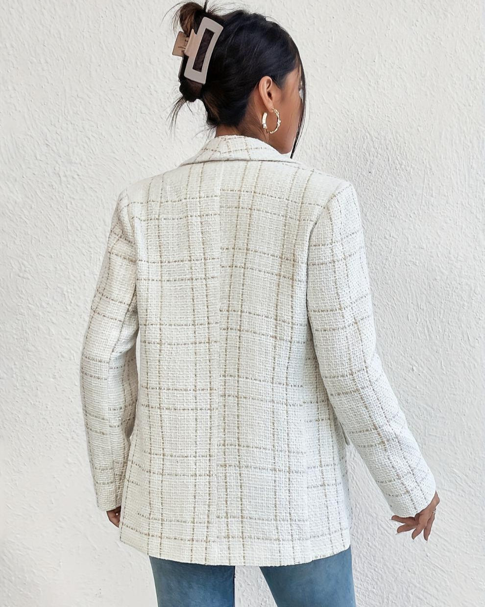 Donna di spalle con blazer a quadri bianco, fermaglio per capelli marrone, orecchini a cerchio, jeans blu. Moda autunnale elegante e casual.