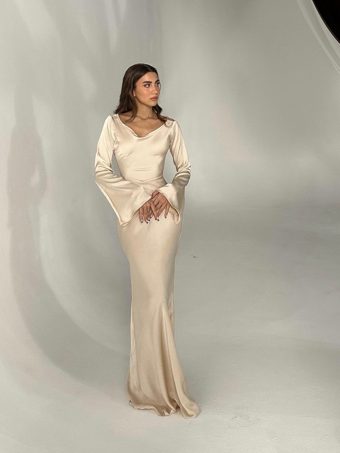 Donna in abito lungo color crema, elegante e aderente, con maniche svasate, posa in studio fotografico minimalista. Moda femminile, abbigliamento elegante.