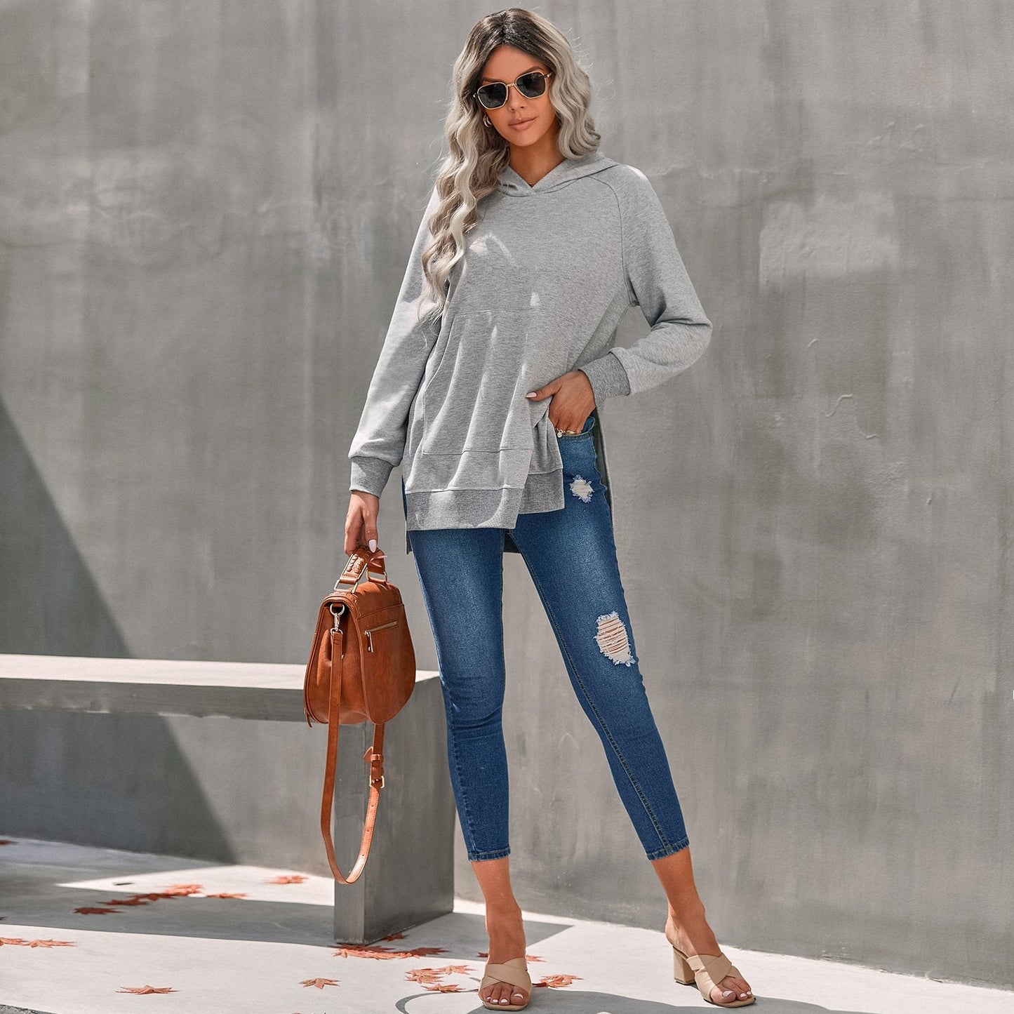 Donna con occhiali da sole indossa felpa grigia e jeans strappati, borsa marrone a tracolla, posa all'aperto. Moda casual autunnale.