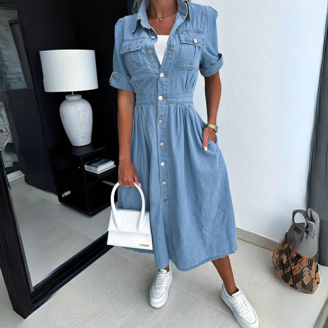 Donna in abito di jeans con bottoni, borsa bianca e sneakers bianche, accanto a specchio e lampada moderna. Moda casual chic.