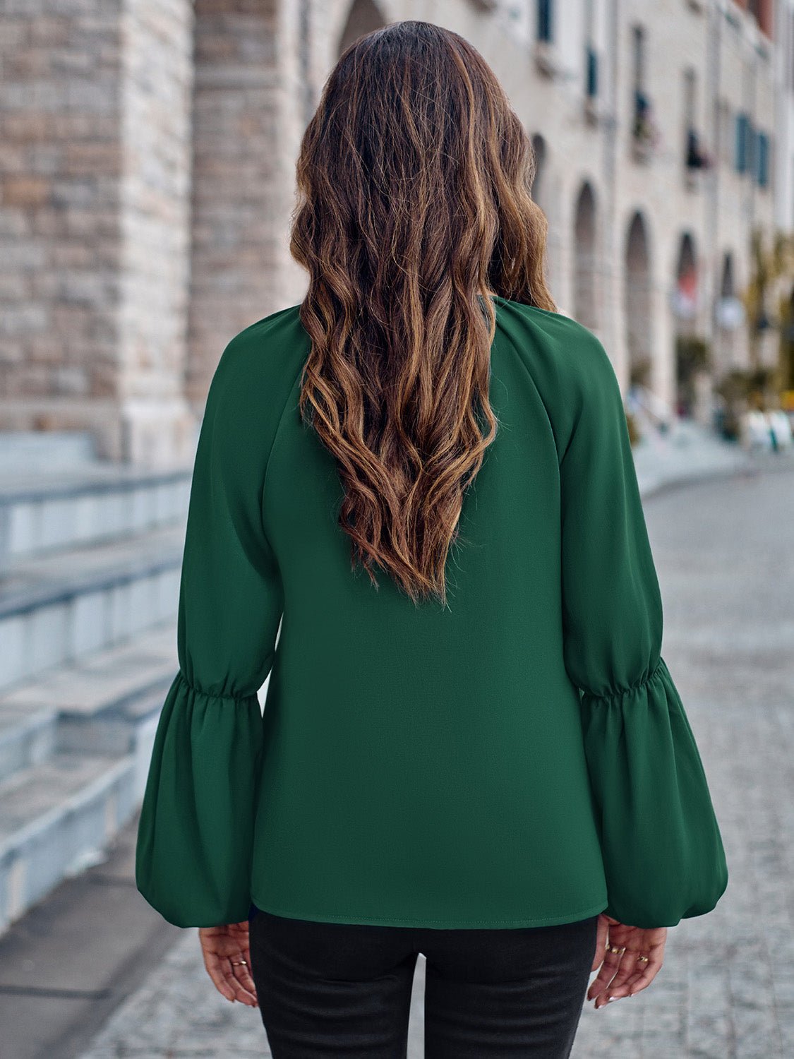 Donna con capelli lunghi ondulati, indossa camicetta verde e pantaloni neri, cammina su strada acciottolata vicino a edifici storici.