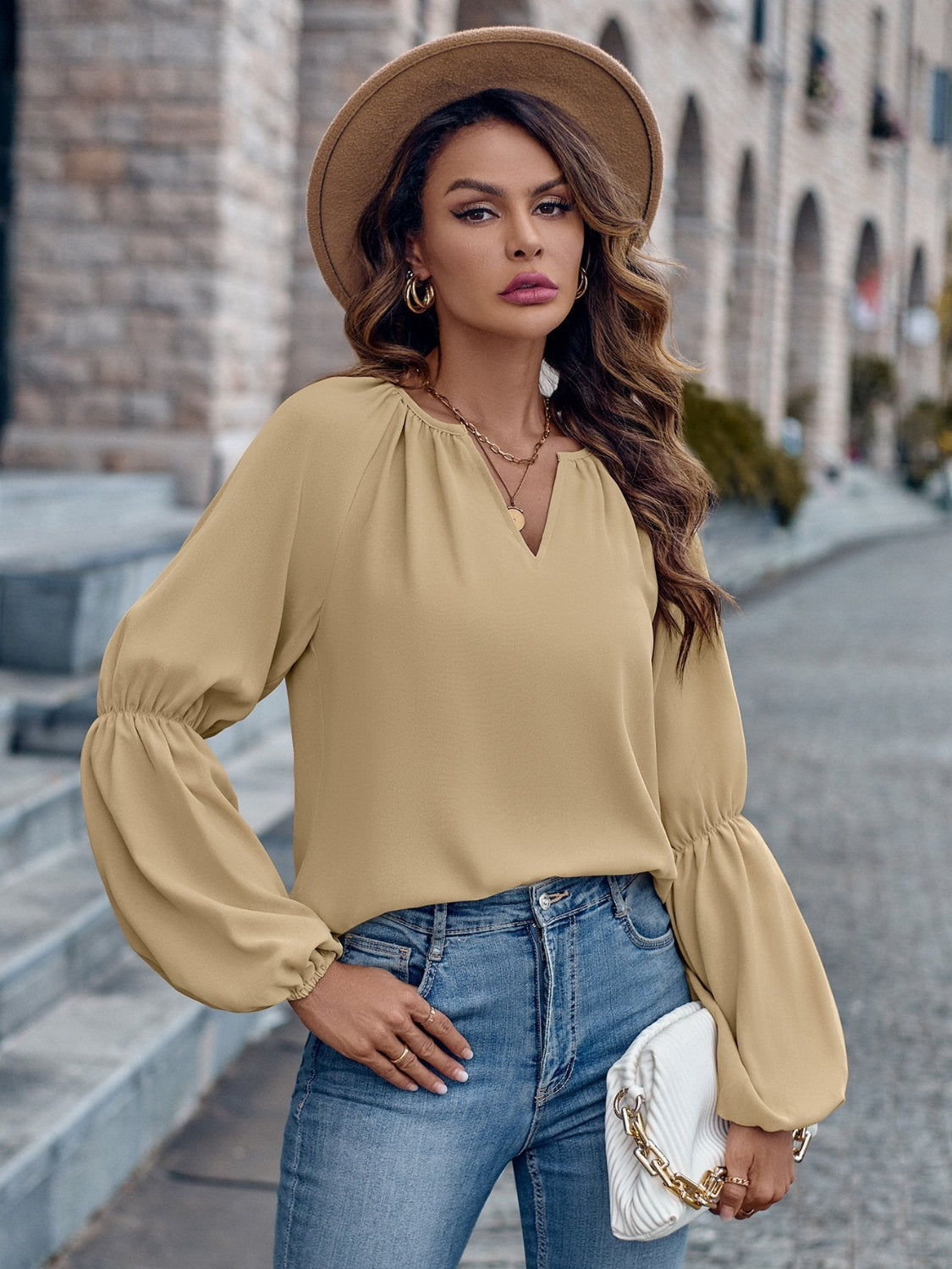 Donna elegante con cappello beige, camicetta a maniche lunghe e jeans, tiene una borsa bianca. Sfondo urbano con edifici in pietra.