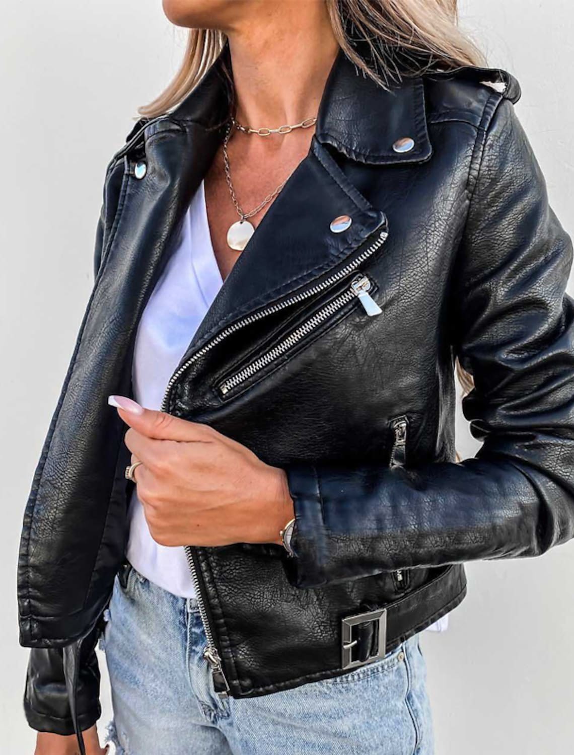 Giacca in pelle nera da donna, stile motociclista, con zip e bottoni argentati, indossata su maglietta bianca e jeans chiari.
