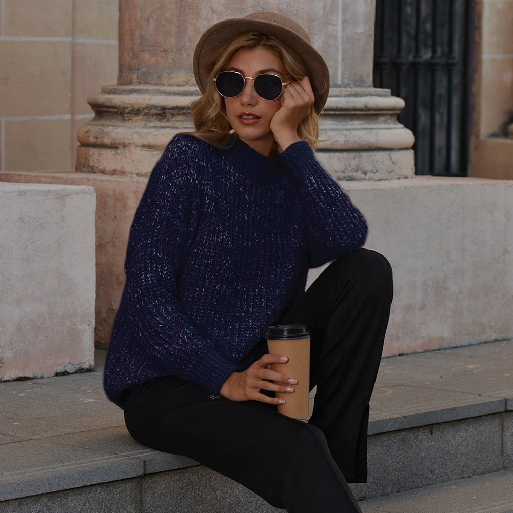 Donna con cappello e occhiali da sole, indossa maglione blu e pantaloni neri, seduta su gradini con caffè in mano. Moda autunnale urbana.