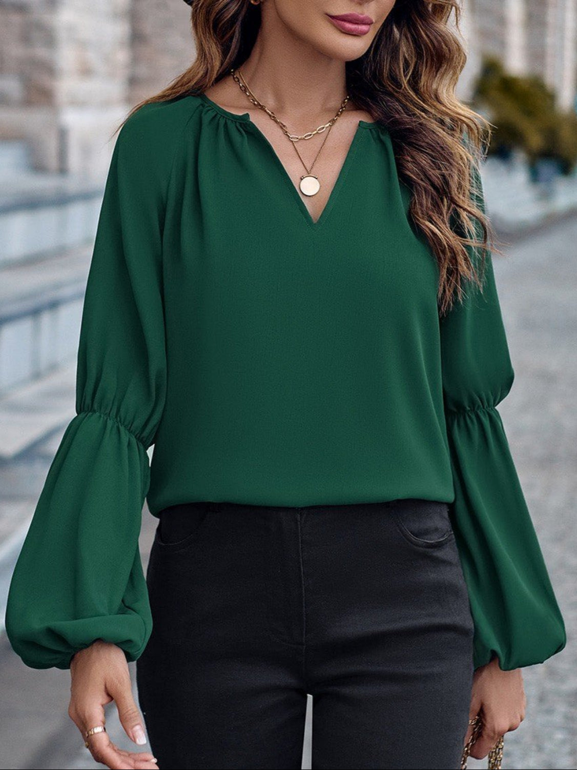 Donna con camicetta verde a maniche lunghe e pantaloni neri, collana dorata, sfondo urbano sfocato. Moda autunnale elegante.
