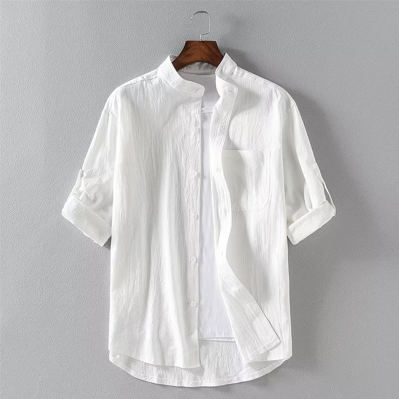 Camicia bianca da uomo in cotone, maniche lunghe arrotolate, colletto alla coreana, appesa su gruccia di legno, sfondo grigio chiaro.