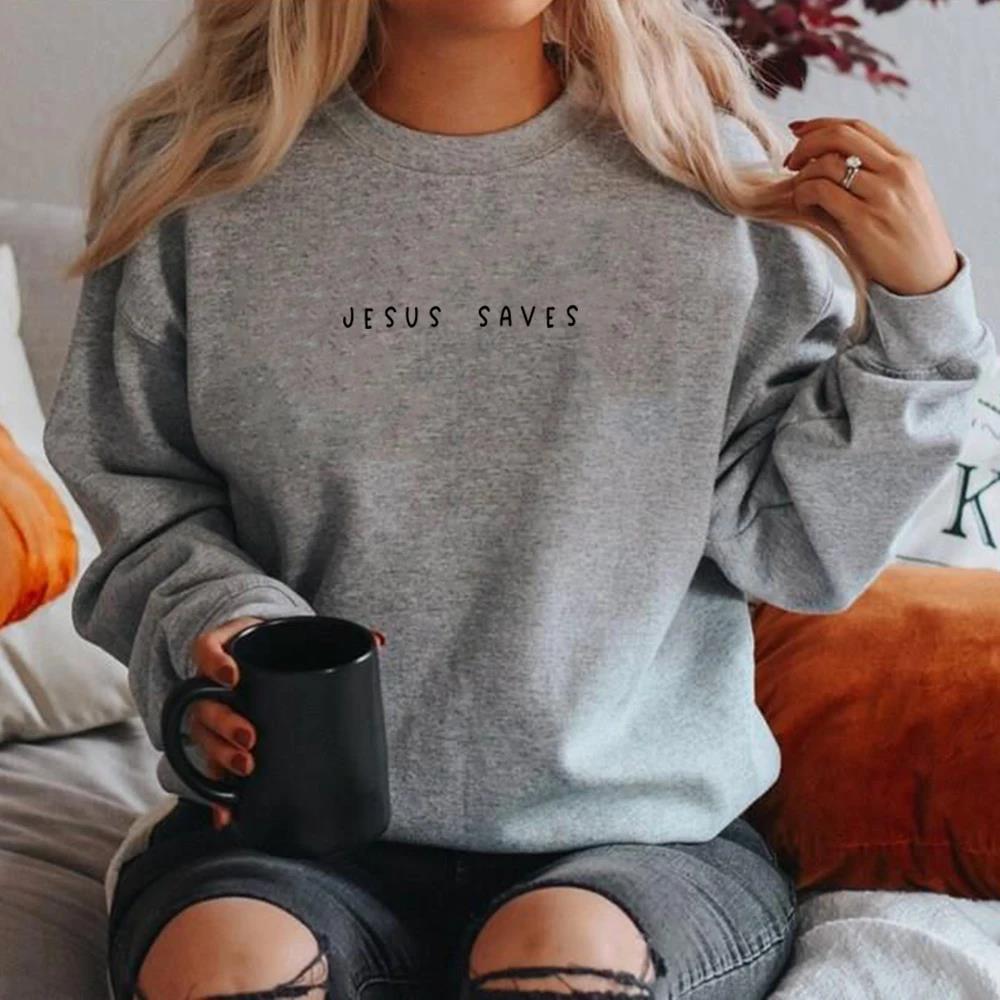Donna con felpa grigia "Jesus Saves", seduta su divano, tiene tazza nera. Moda casual autunnale, look accogliente, abbigliamento religioso.