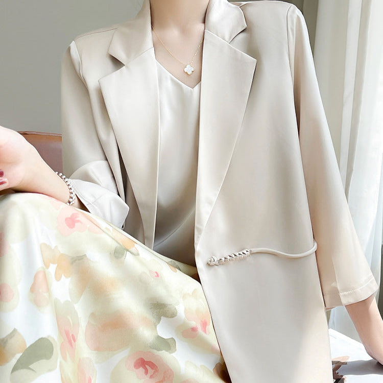 Donna in abito elegante beige, giacca sartoriale e collana con ciondolo, seduta su sedia. Moda femminile, abbigliamento chic, stile sofisticato.