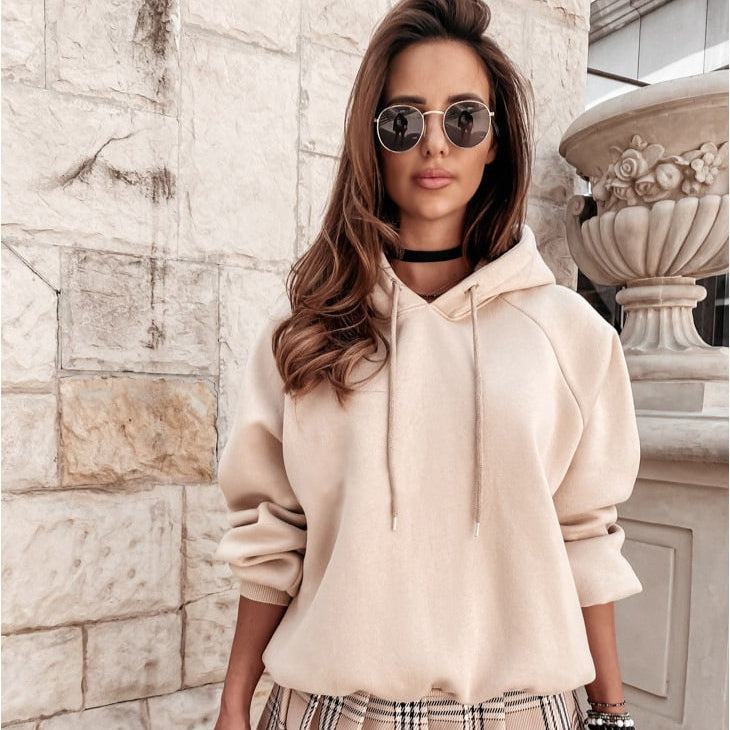 Donna con occhiali da sole indossa felpa beige e gonna a quadri, posa davanti a muro di pietra. Moda autunnale, stile casual chic.