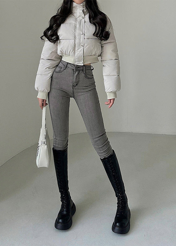 Donna in giacca imbottita bianca, jeans grigi aderenti e stivali neri alti. Moda invernale elegante, outfit casual chic, borsa bianca a mano.