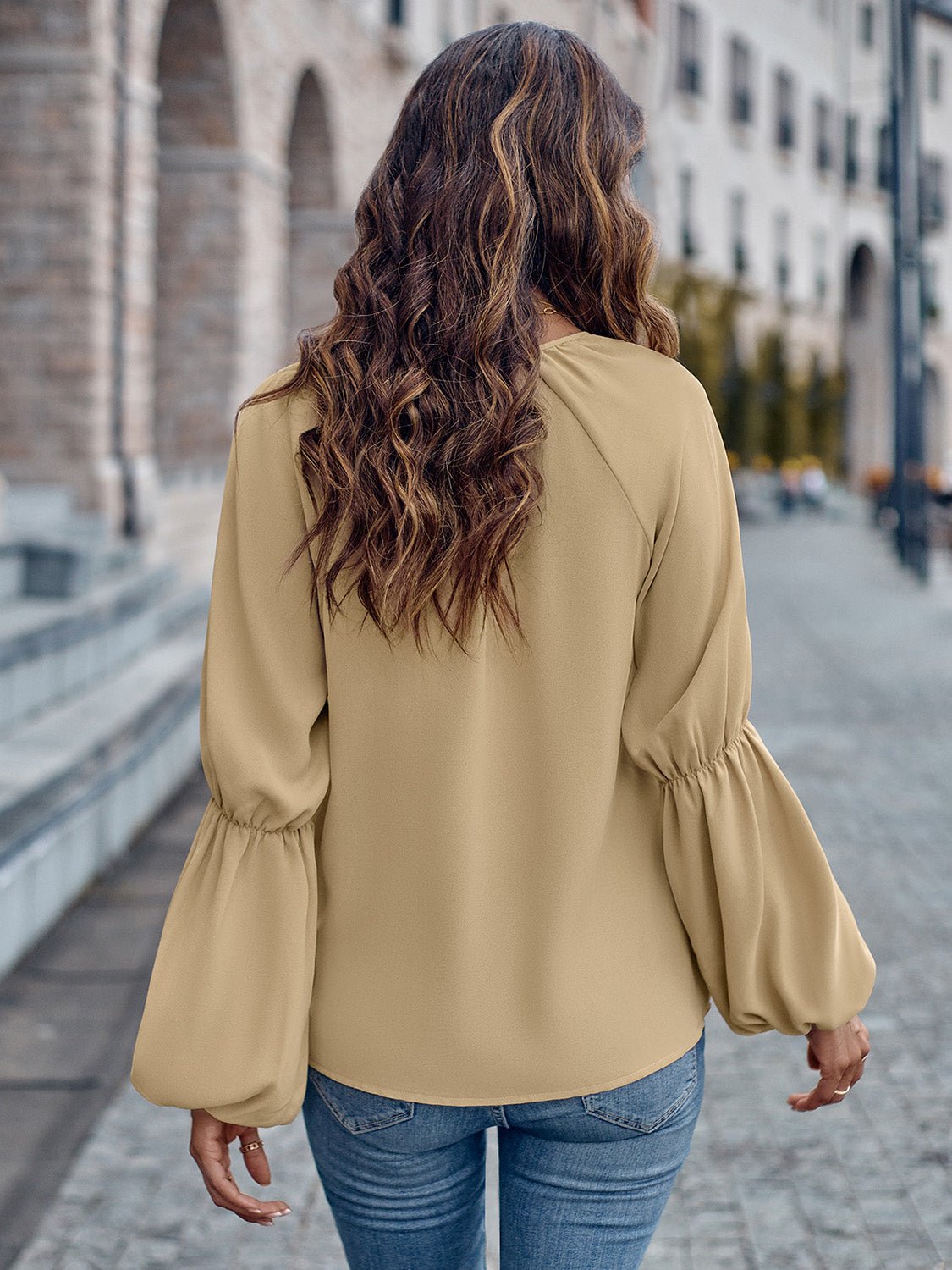 Donna di spalle con capelli mossi, indossa una camicetta beige a maniche lunghe e jeans, cammina su un marciapiede cittadino.