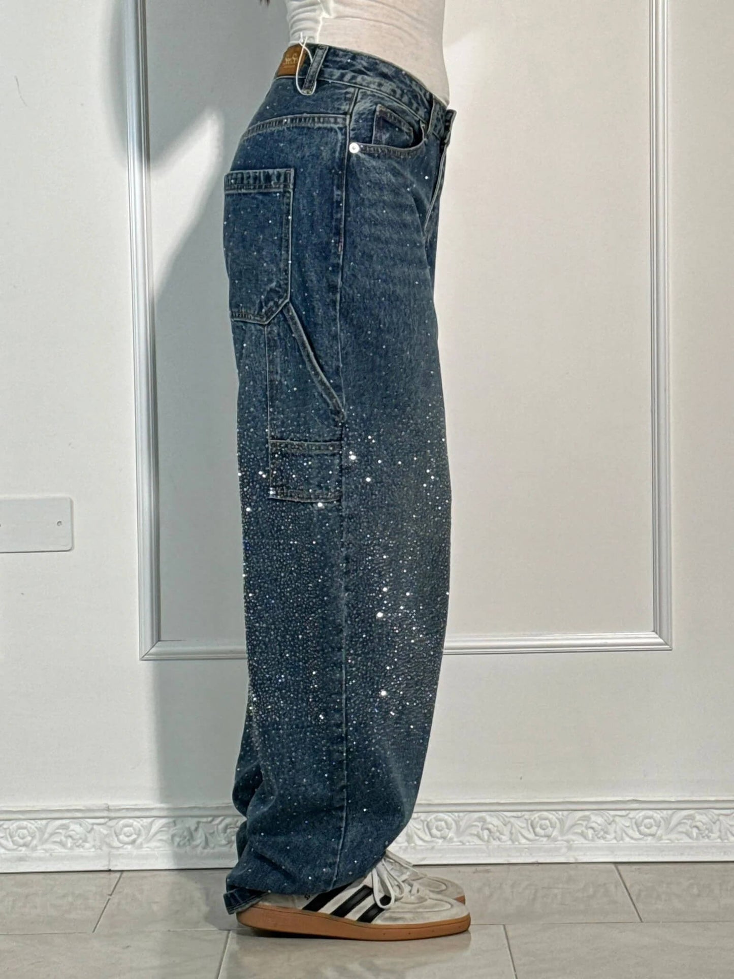 Jeans larghi in denim blu con paillettes scintillanti, tasche cargo, indossati con sneakers bianche e nere, su sfondo bianco minimalista.