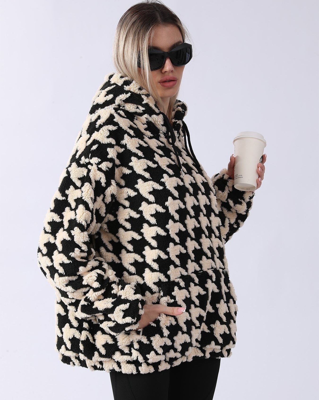 Donna con occhiali da sole indossa felpa oversize in pile con motivo pied de poule bianco e nero, tiene bicchiere da caffè. Moda invernale casual.