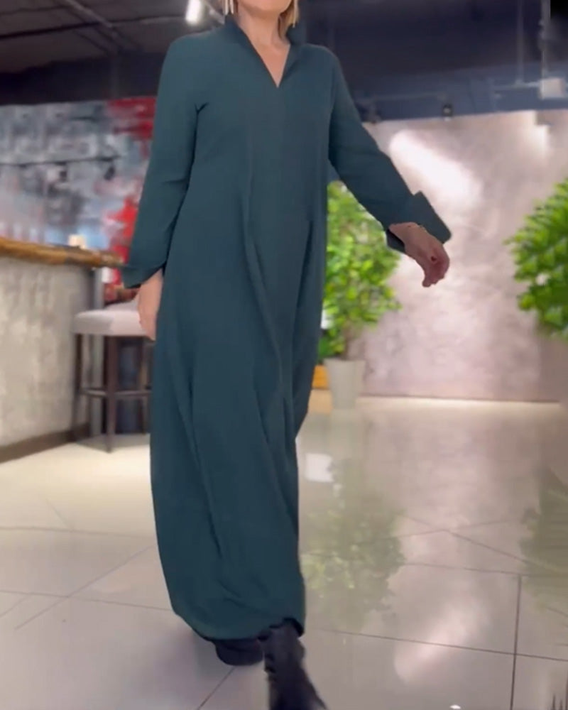 Donna in abito lungo verde scuro cammina in un ambiente moderno con piante e arredi eleganti. Moda femminile, abbigliamento casual chic.