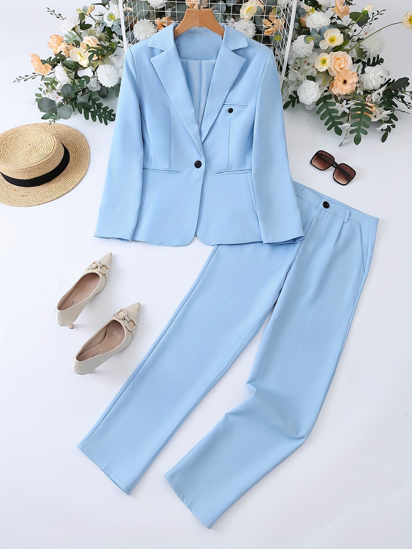 Completo elegante donna azzurro, giacca e pantaloni, con scarpe beige, cappello di paglia e occhiali da sole, su sfondo floreale. Moda femminile.