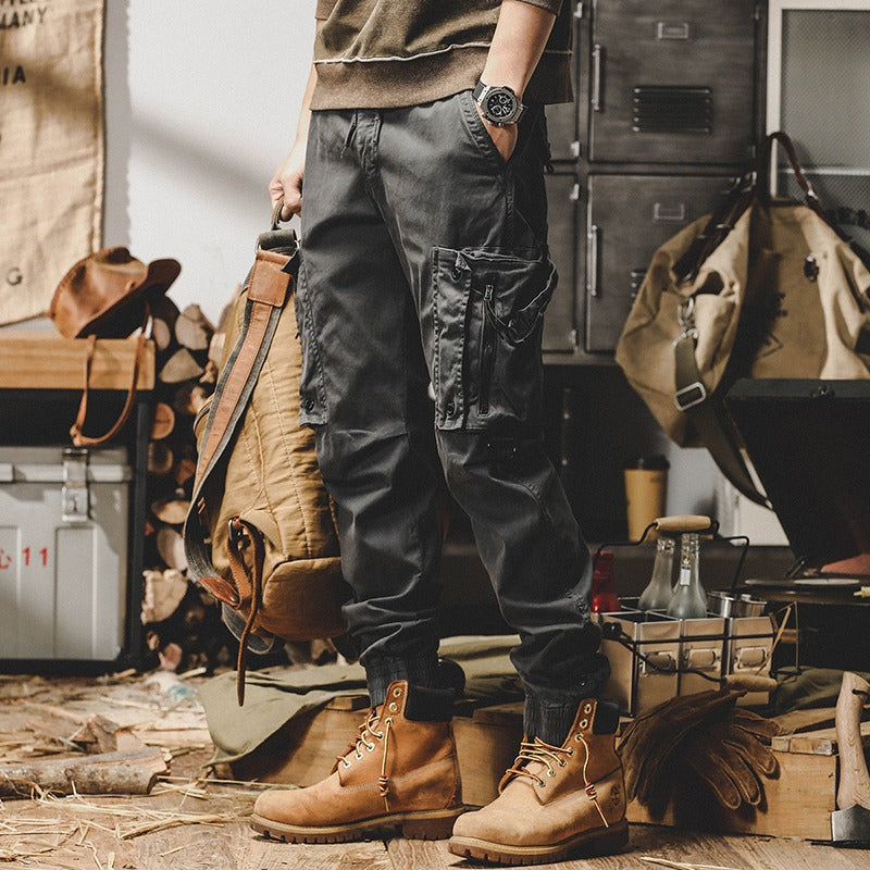 Uomo con pantaloni cargo neri e stivali marroni in una stanza rustica, circondato da attrezzi e zaini. Stile casual, moda outdoor.