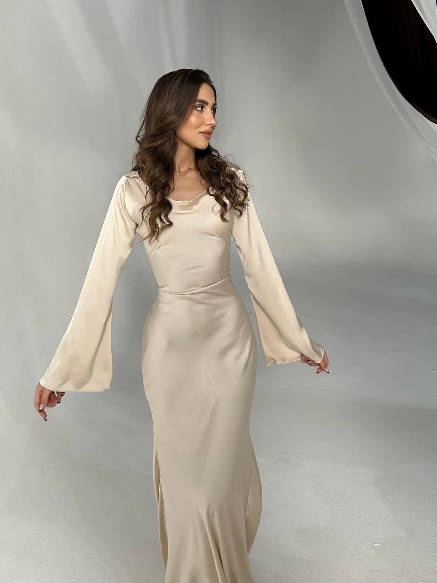 Donna in abito lungo color crema con maniche ampie, posa su sfondo bianco. Moda elegante, abbigliamento femminile, stile sofisticato.