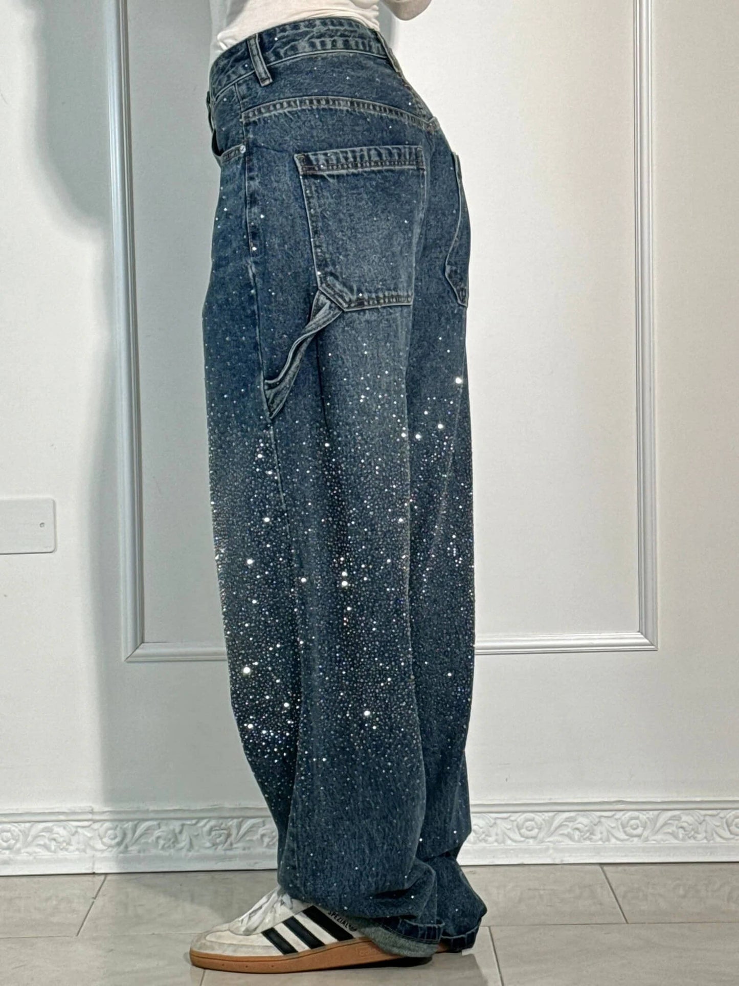 Jeans larghi in denim blu con dettagli scintillanti, tasche laterali, abbinati a scarpe da ginnastica bianche con strisce nere, su sfondo neutro.