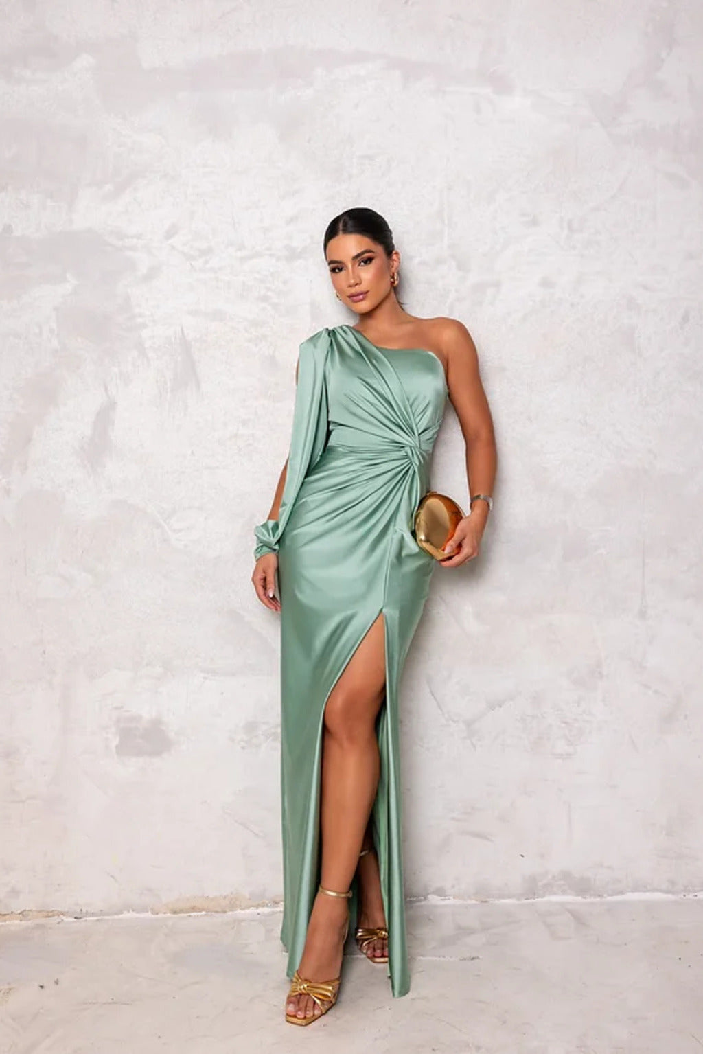 Donna in abito verde elegante monospalla con spacco alto, tiene una pochette dorata. Moda serale, abbigliamento da cerimonia, stile glamour.