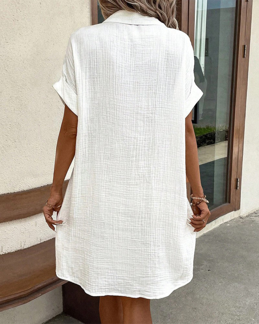 Donna con abito camicia bianco in cotone, maniche corte, davanti a una porta. Moda estiva, abbigliamento casual, stile minimalista.