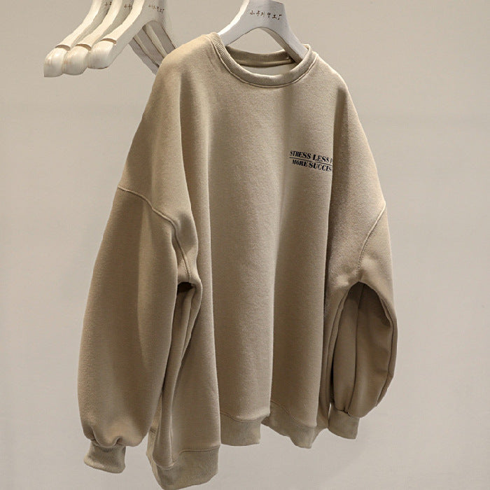Felpa beige oversize con scritta "STRESS LESS MAKE SUCCESS", appesa su gruccia bianca. Moda casual, abbigliamento comodo, tendenze streetwear.