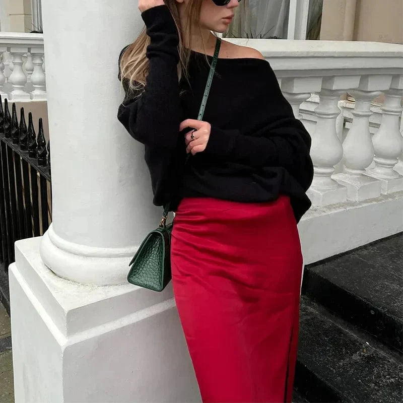 Donna con maglione nero e gonna rossa, borsa verde a tracolla, appoggiata a una ringhiera bianca. Moda autunnale elegante e casual.